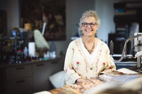 Anna Chupa stands in her studio and smiles at the camera for a portrait. Before her in a table with a quilt in progress.